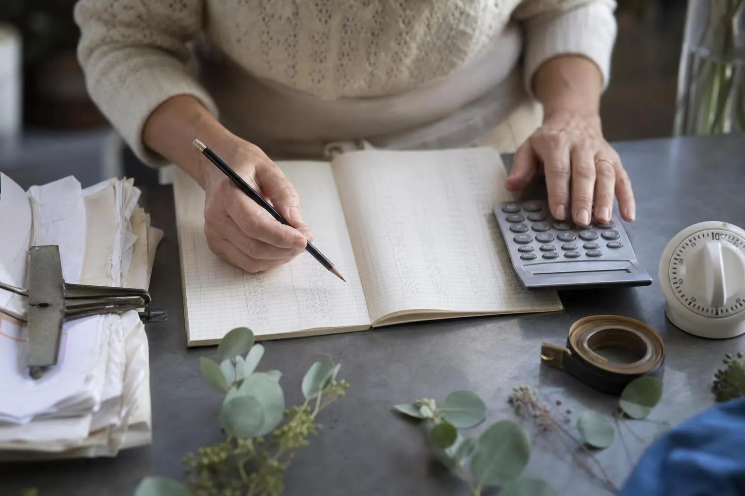 Gros plan sur les mains d’une fleuriste additionnant des lignes de bordereau au crayon à côté d’une calculatrice de bureau et d’un minuteur